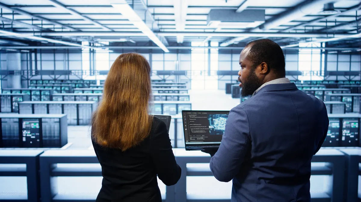 Two engineers reviewing code on a laptop while standing in a data center aisle lined with server racks