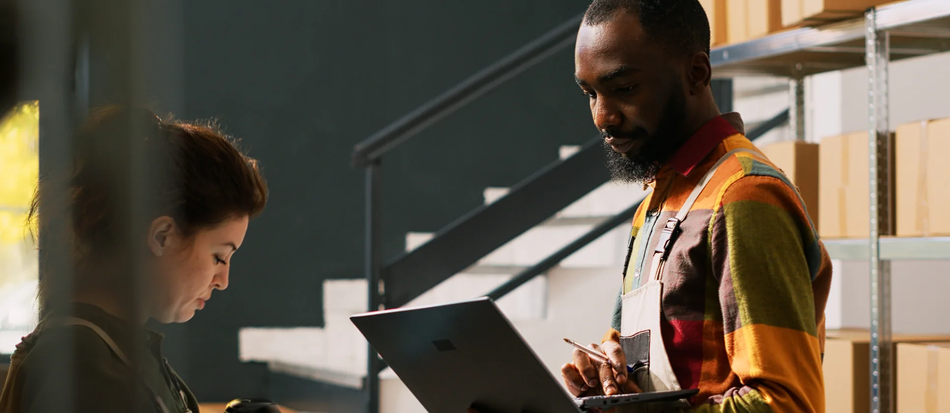 Warehouse fulfillment worker reviewing checkout on a laptop while a colleague scans inventory nearby