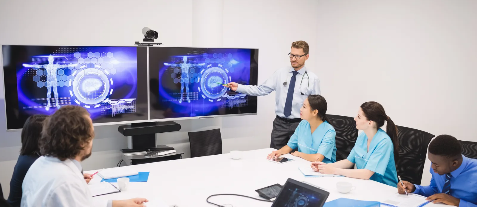 Doctor presenting patient scan data to a clinical team on large screens in a hospital meeting room